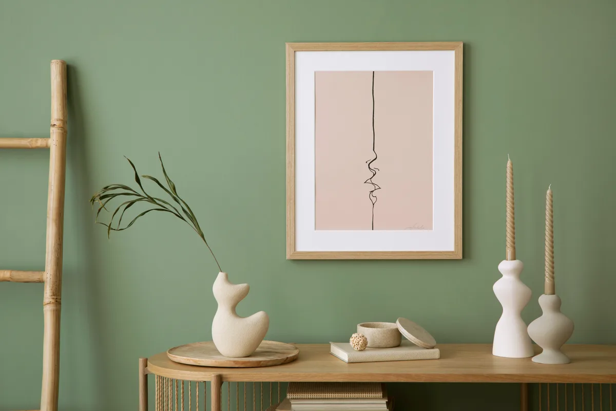 Als Akzentfarbe passen erdige Farbtöne wie Salbeigrün sehr gut. Eine salbeigrüne Wand in einem Wohnzimmer mit verschiedenen Holzmöbeln und einem Bild davor.